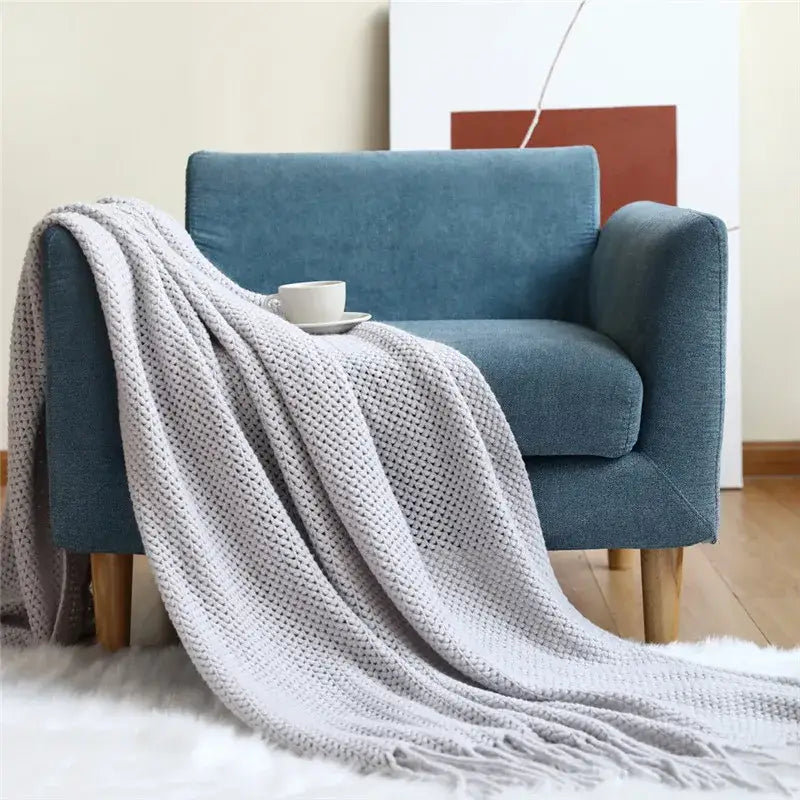 A light gray cozy nordic knit textured throw blanket with fringed edges draped over a blue armchair for nordic knit sofa decorbites cozy nordic