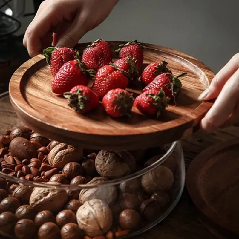 A round wooden serving plate with natural finish holds red strawberries alongside 3 tier glass food storage containers with wood stand