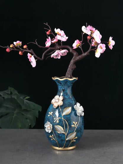 A blue ceramic vase with gold accents and floral embellishments holds dried flowers on a wobble plate display