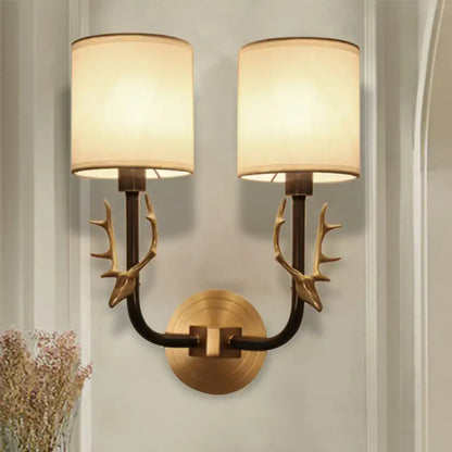 Antique brass antler wall sconce featuring stylish design with brass-toned arms and two cylindrical cream fabric shades for bedroom lighting