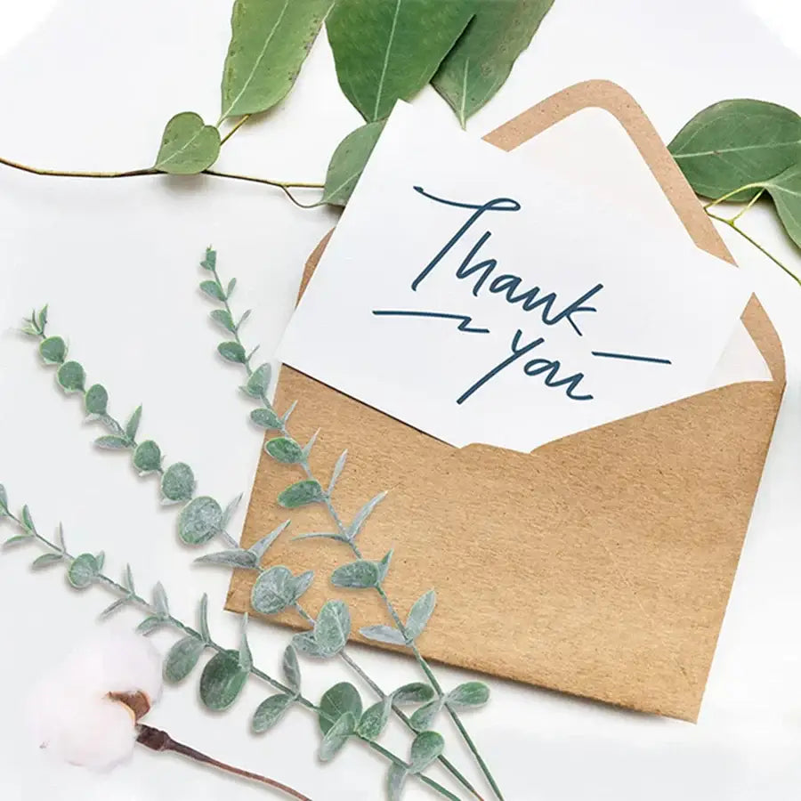A rustic brown kraft paper envelope holding a white thank you card alongside artificial eucalyptus greenery stems for living room decor