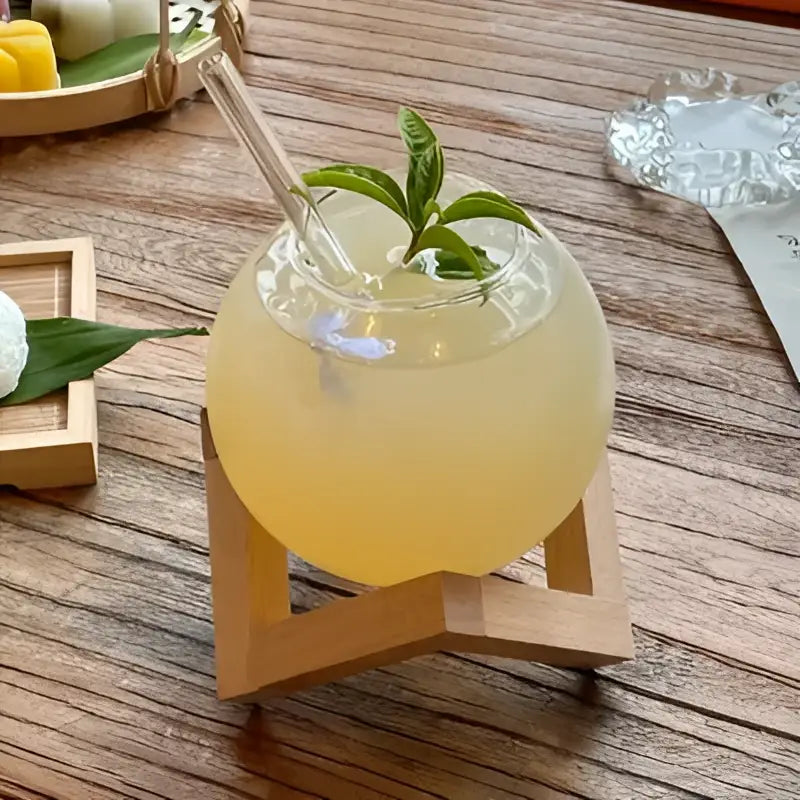 A clear glass ball shape coffee juice cocktail glass with wooden stand filled with pale yellow drink and mint garnish