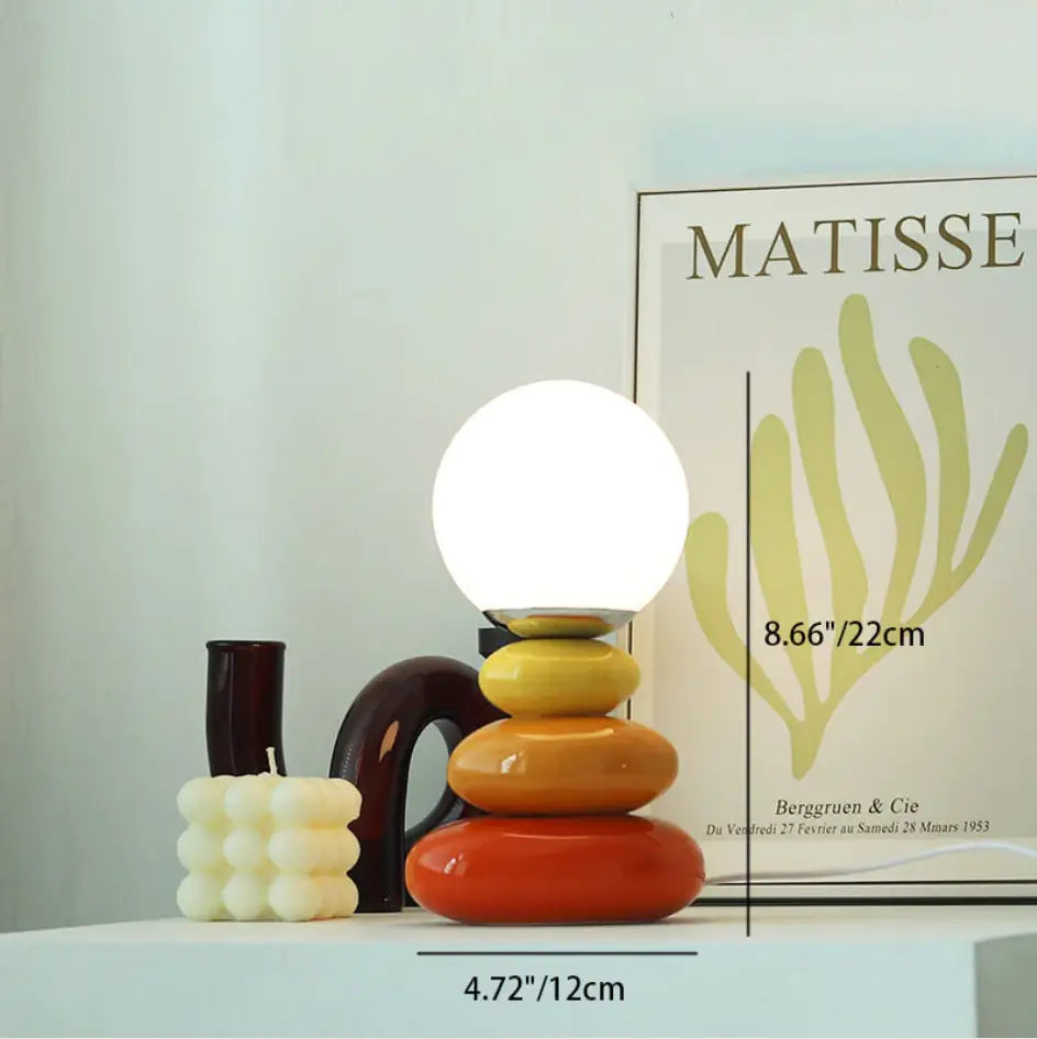 A bauhaus table lamp with a vibrant orange and yellow ceramic tiered base and a glowing white spherical glass shade