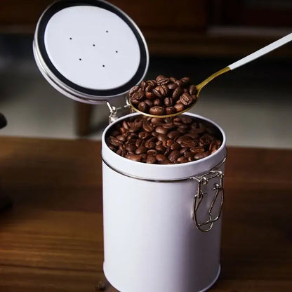 White cylindrical metal coffee canister with silver lid and clasp filled with dark roasted beans for hermetic food storage
