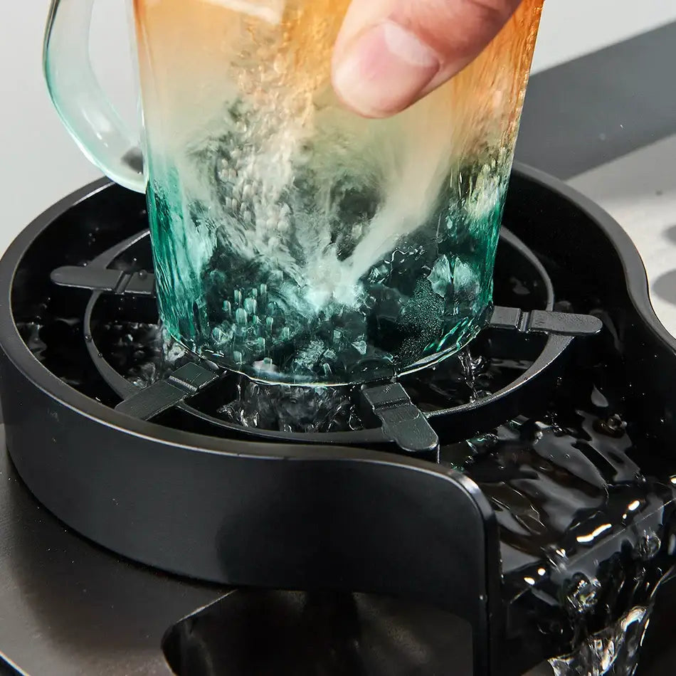 A gradient-colored glass mug with orange to teal textured surface near a black plastic glass rinser faucet accessory