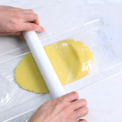 A white rolling pin is used to flatten yellow dough wrapped in plastic alongside a blue silicone rolling balance ruler