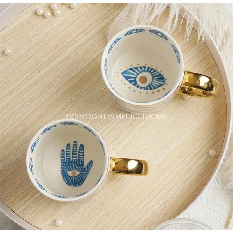 Two ceramic mugs with gold-toned handles featuring blue hamsa hand and blue eye motifs from decorbites bright eyes collection