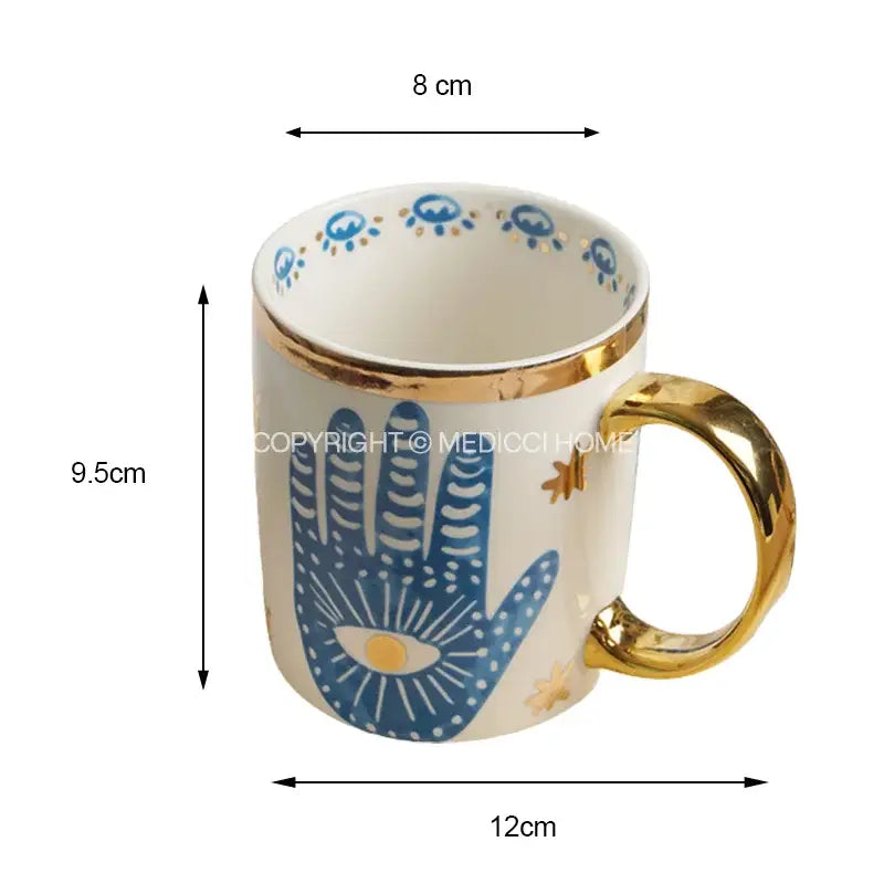 A white ceramic mug with gold rim and handle featuring a blue hamsa hand eye design for bright eyes ceramic decorbites