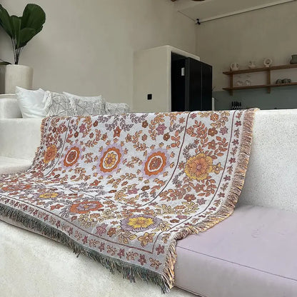 A cozy, off-white woven throw blanket featuring a vibrant floral pattern in shades of orange, yellow, and muted purple, with decorative fringe along its edges.