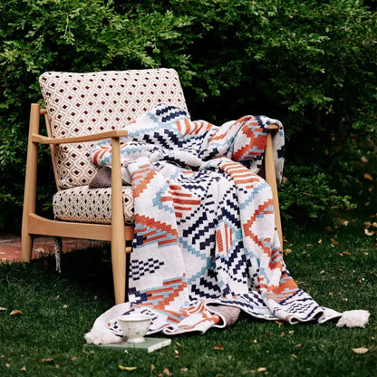 A cozy hand-knitted boho geometric summer blanket in orange navy blue and teal draped over a wooden armchair