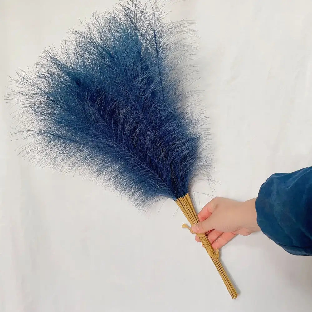 A hand holds a bundle of deep blue fluffy pampas grass with natural bamboo handles for living room decor