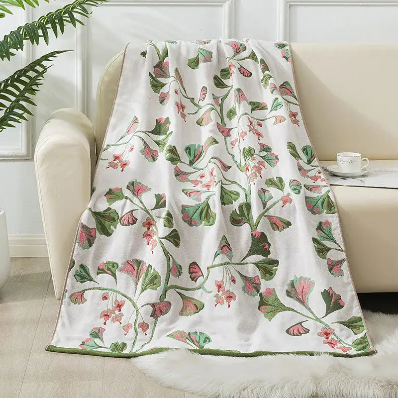 A soft white decorbites cotton gauze bath towel with pink and green ginkgo leaf pattern draped over a cream sofa