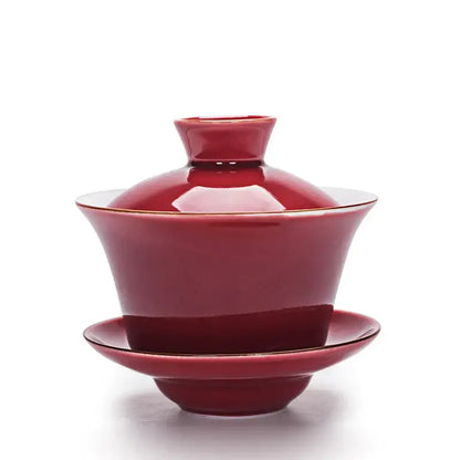 A glossy crimson ceramic gaiwan with gold trim complementing a celadon tea set with gold rim tea tureen
