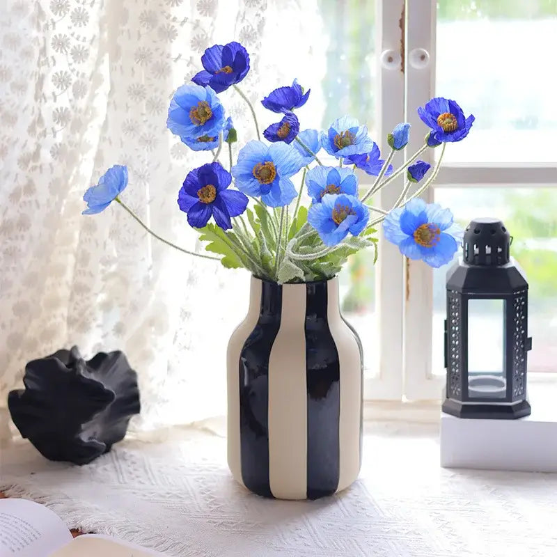 A sleek black and cream striped ceramic vase holding vibrant blue artificial poppies in a striped ceramic vase design