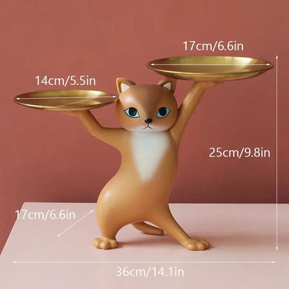 A tan resin cat figurine with blue eyes balancing golden plates in a ceramic white and beige cat figurines storage tray