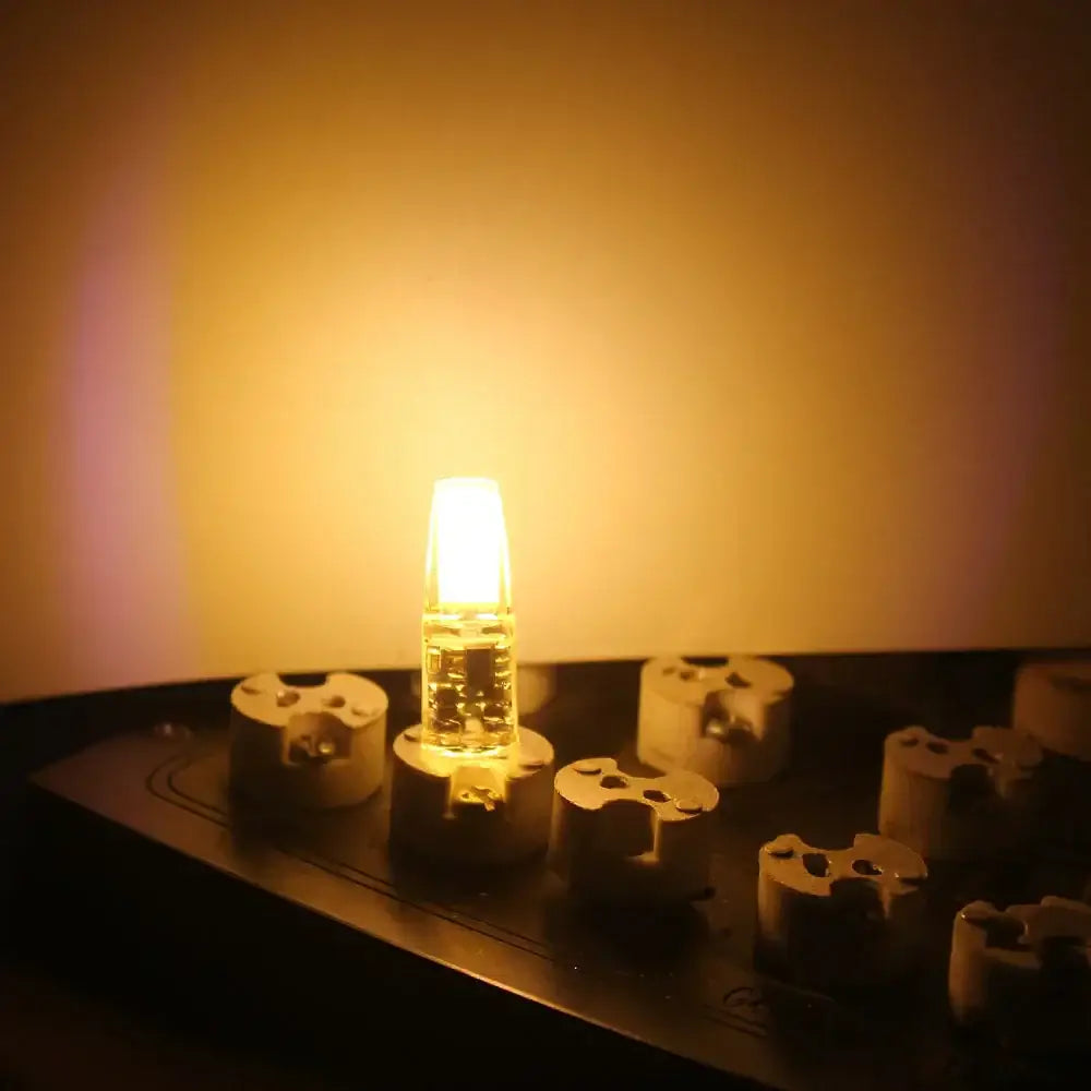 A glowing amber dimmable g4 12v led bulb with clear glass housing and metallic base atop beige ceramic insulators