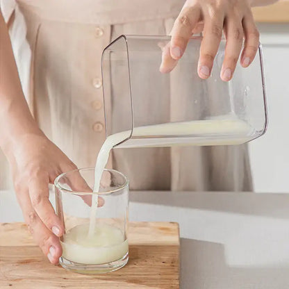 Clear rectangular glass pitcher pouring milk into glass next to clear greek yogurt maker with white lid and mesh filter