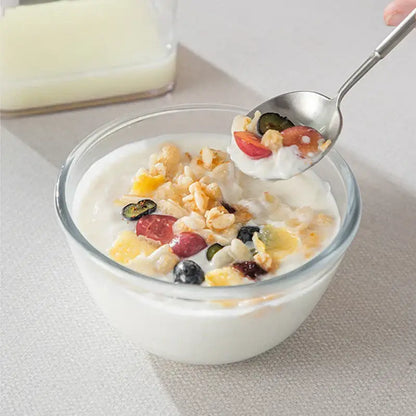 A clear glass bowl with creamy greek yogurt topped with mixed fruit and granola beside a clear greek yogurt maker with white lid and mesh filter