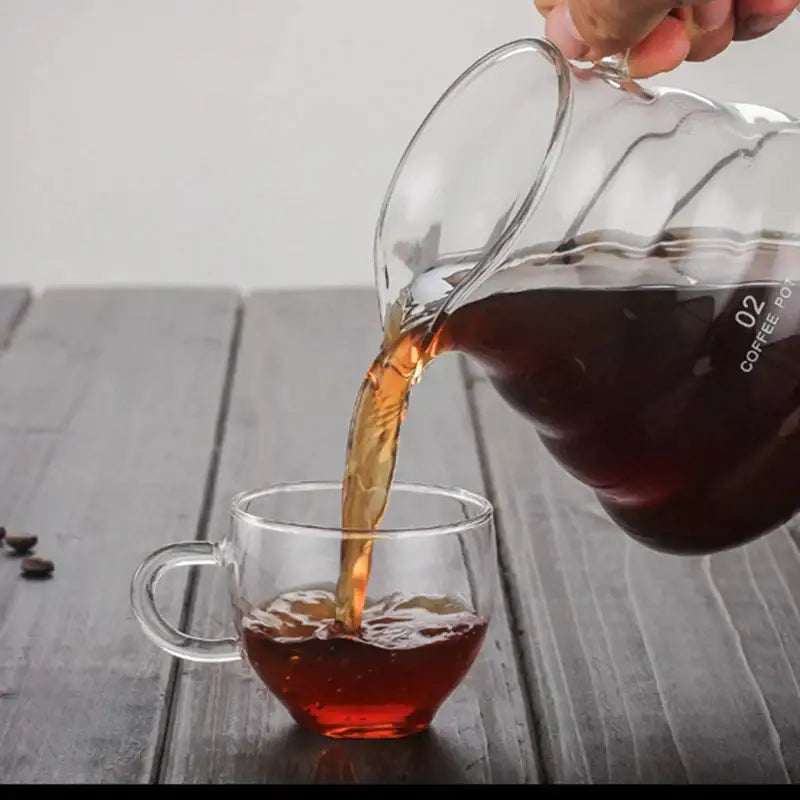 Clear glass cloud shaped glass coffee kettle with wavy design pouring dark espresso into matching clear glass mug