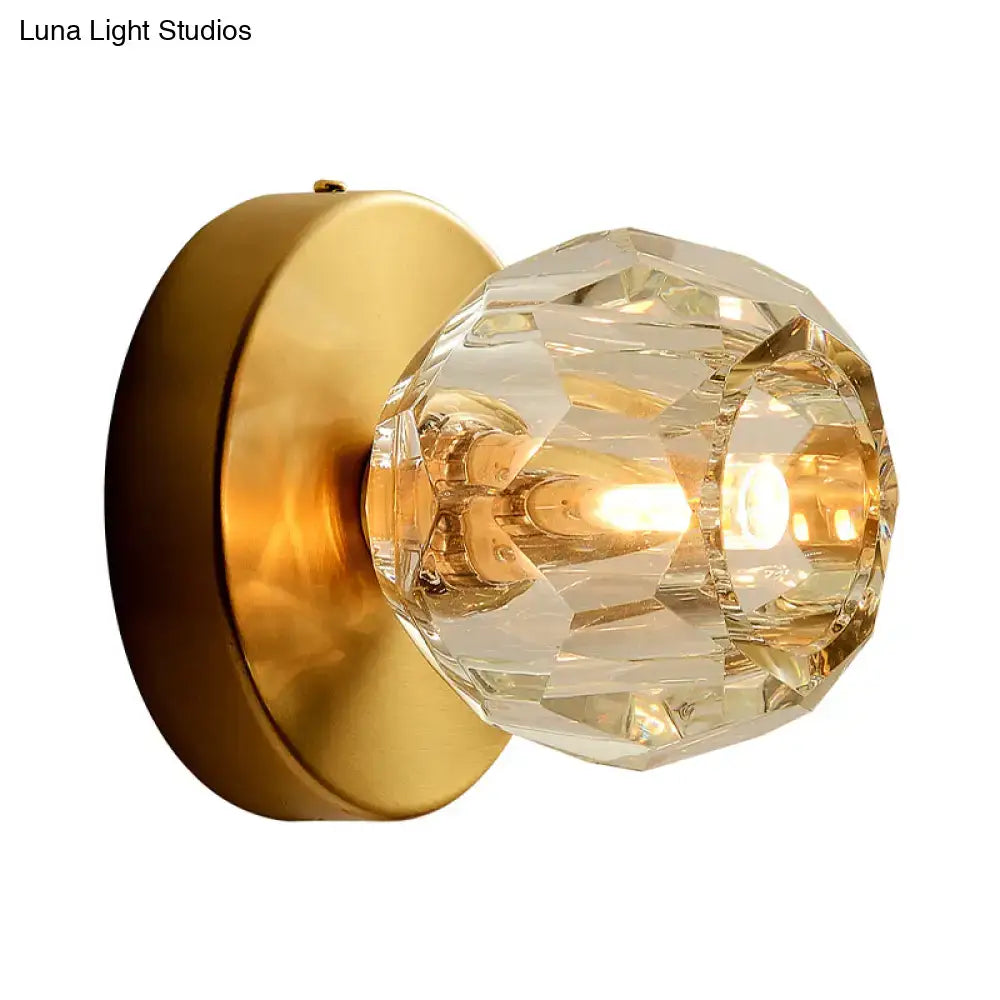 Contemporary golden wall mount lamp featuring a faceted glass shade with a warm light bulb on a brass plate