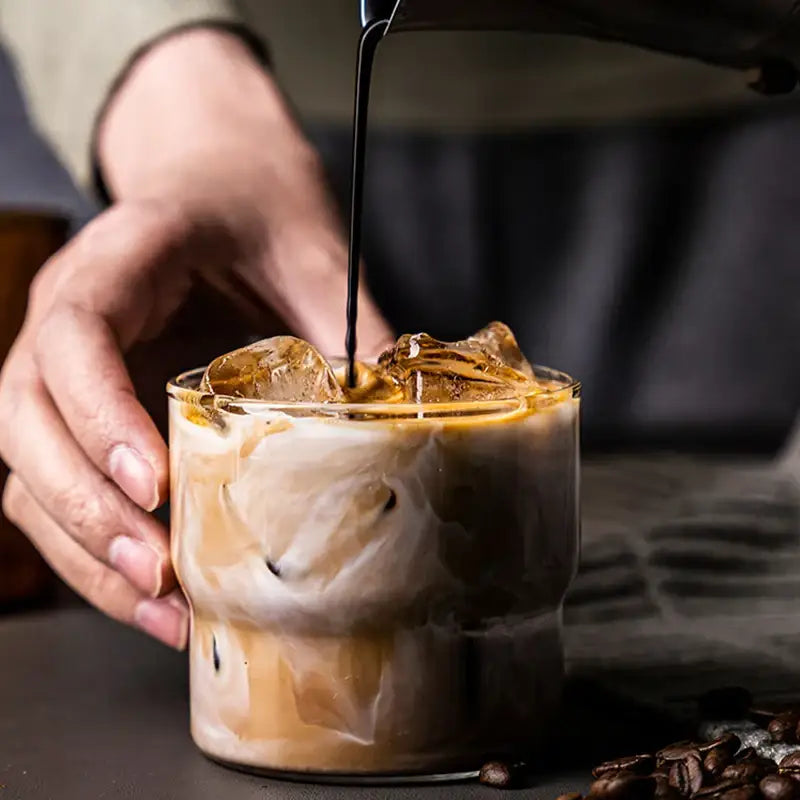 A hand holds a clear decorbites creative glass coffee smoothie cup with iced coffee layered with espresso and milk