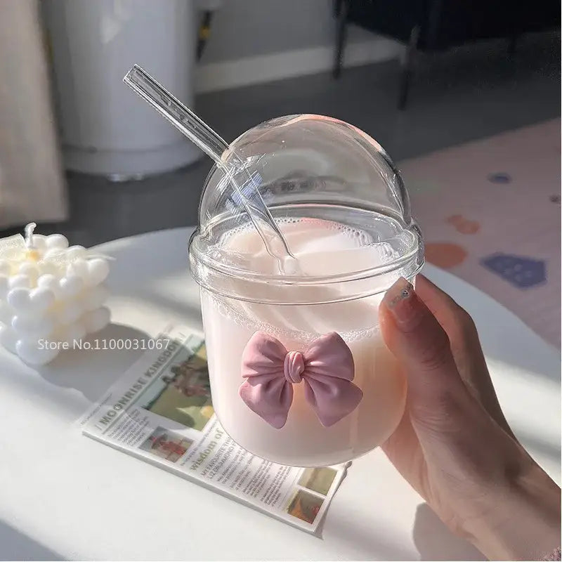 A clear glass cup with a dome lid decorated with a pink bow tie for kitchen coffee tumbler use