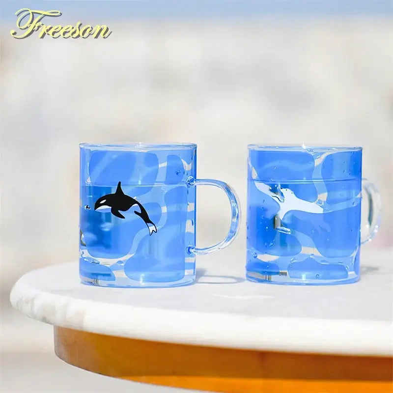 Two translucent blue glass coffee mugs with orographic whale designs and heat resistant handles for kitchen use