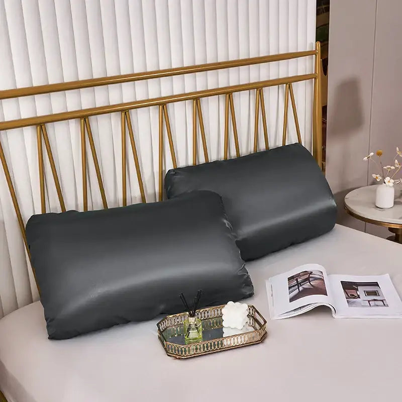 Two dark gray decorbites imitation silk pillowcases with elastic band rest on a bed with a gold headboard