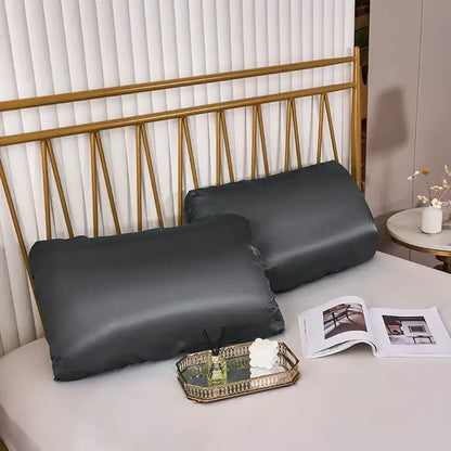 Two dark gray decorbites imitation silk pillowcases with elastic band rest on a bed with a gold headboard