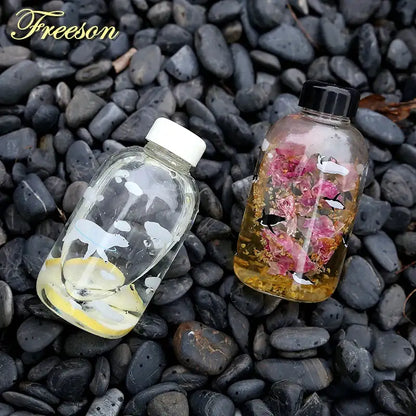 Two transparent glass water bottles with white whale silhouettes lemon slices and pink roses with black birds on dark pebbles