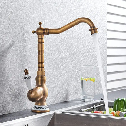 Antique brass kitchen sink faucet with white ceramic handle and black ornate detailing for hot and cold water mixer