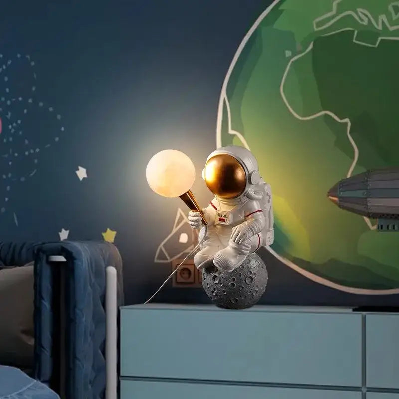 DecorBites™ astronaut led table lamp in white resin with gold helmet seated on textured gray moon surface holding glowing lamp