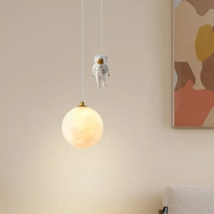 A whimsical white astronaut figurine hangs above a glowing textured moon pendant light from decorbites astronaut moon