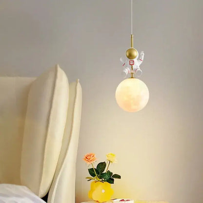 A whimsical white pendant light with gold accents from decorbites astronaut moon hangs above a glowing bedside globe