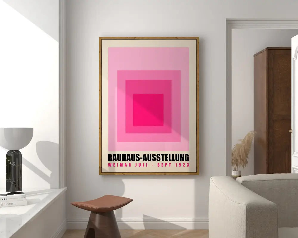 A bright geometric canvas print featuring pink concentric squares and black bauhaus text in a light wood frame