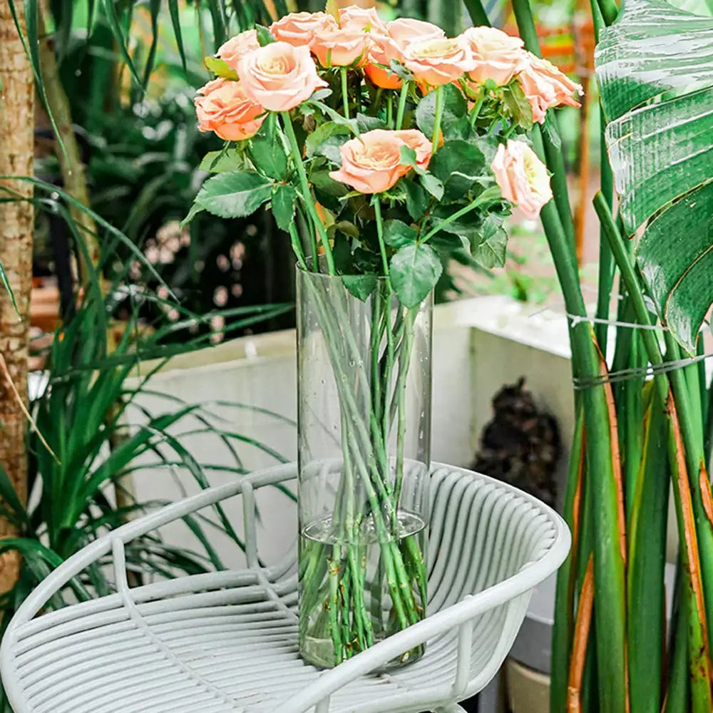 A large clear acrylic hurricane flower vase by DecorBites holding peach-colored roses with green stems and leaves