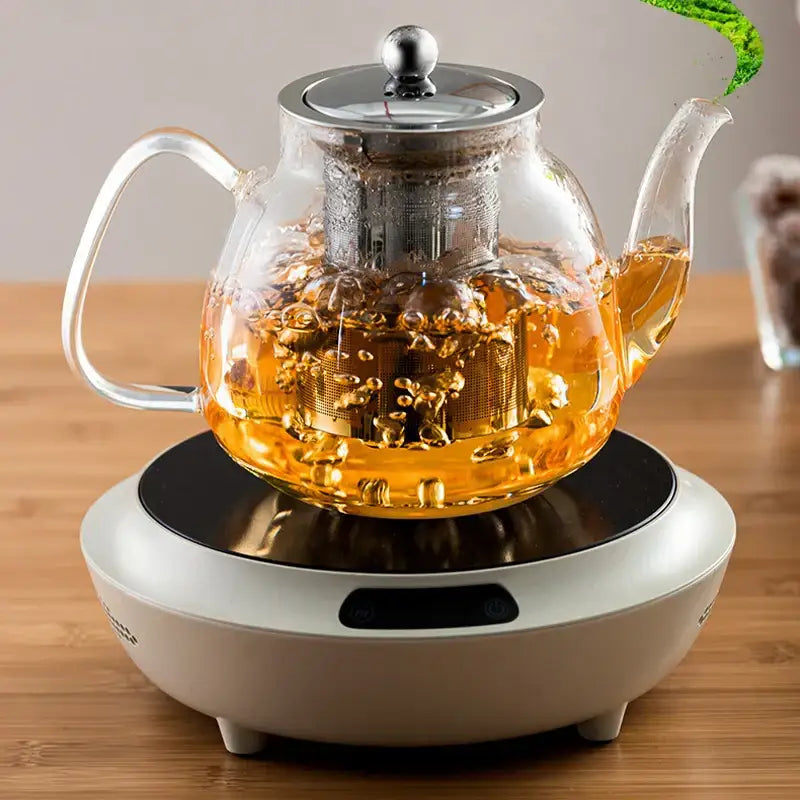 DecorBites glass tea pot with stainless steel filter filled with steaming golden tea on white electric heating base