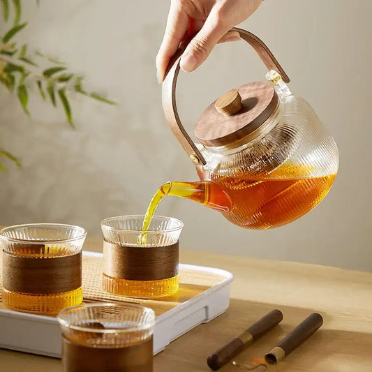 Clear glass tea pot with wooden handle and lid pouring amber tea into matching glasses decorbites glass tea set