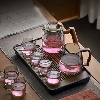 Clear glass decorbites glass tea pot with wooden handle and lid on black tray with glasses filled with pink tea