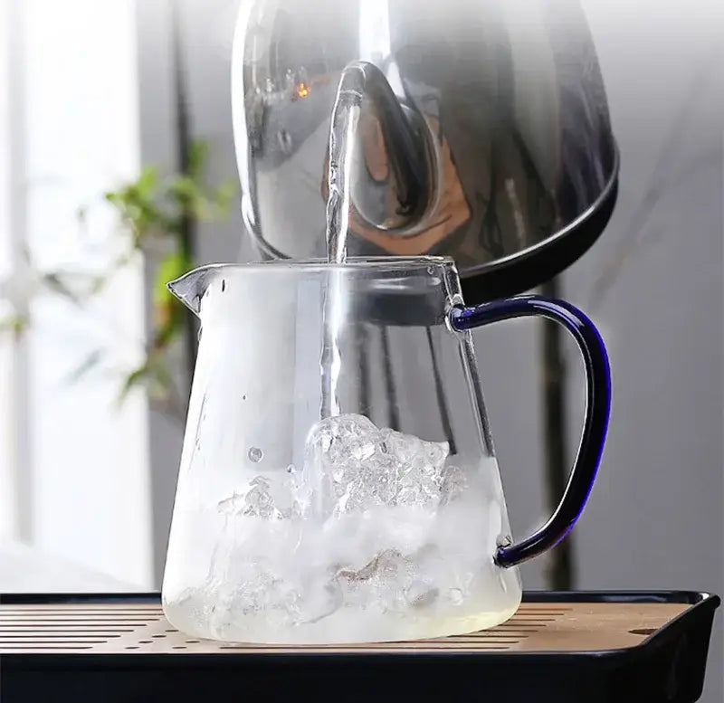 A decorbites glass teapot 550ml with a blue handle is being filled with hot water from a stainless steel kettle