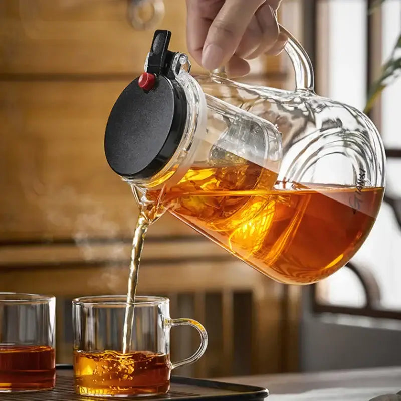 Clear glass decorbites glass teapot with black lid and red knob pouring amber tea using one-button filter technology