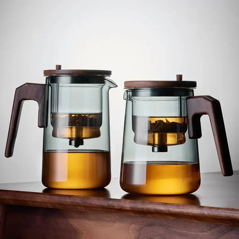 Two decorbites glass teapot infusers with dark wood handles and metal strainers containing loose tea leaves