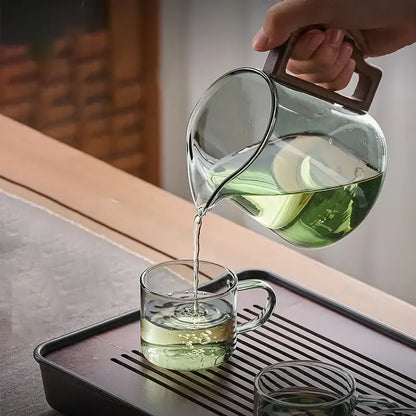 Clear glass decorbites glass teapot with dark brown wood handle pouring green tea into matching glass cup for kung fu tea ceremony