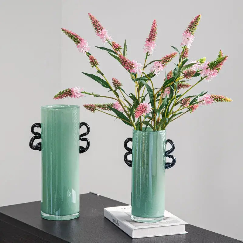 A sleek mint-green decorbites glass vase with glossy finish and black handles holding pink flowering stems