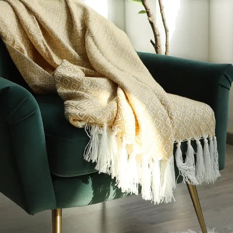 A decorbites golden knitted cream geometric throw blanket with white fringe draped over a dark green velvet chair