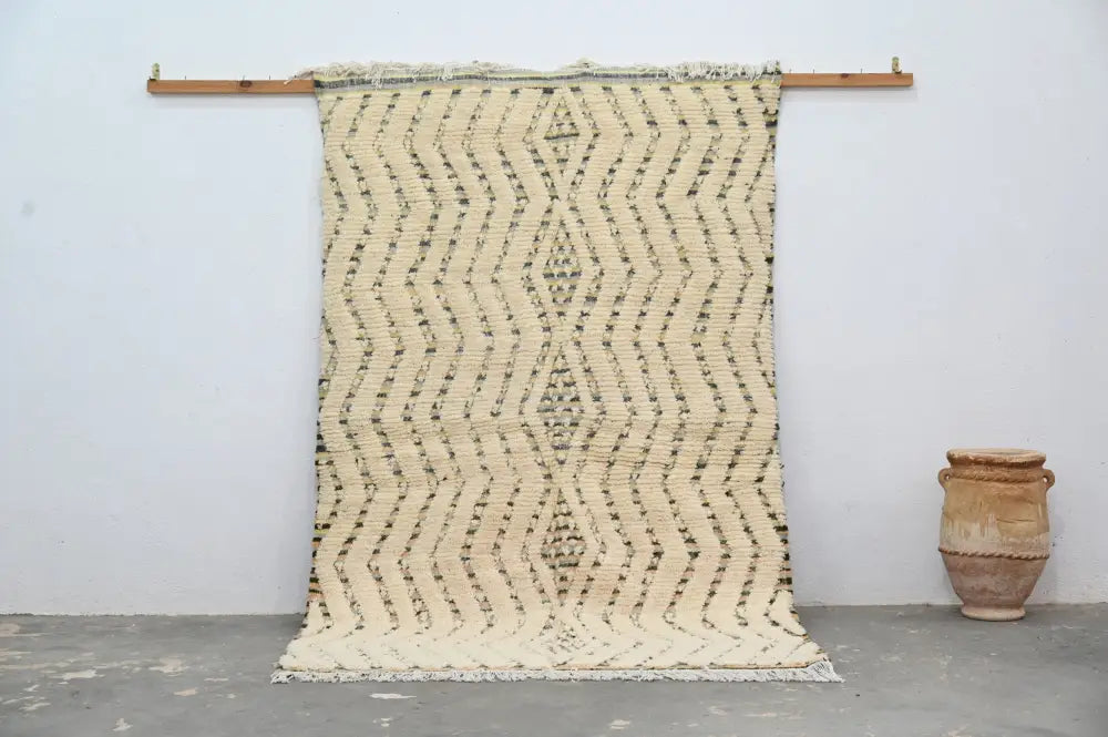 Cream-colored handmade moroccan azilal rug 240x155cm with dark brown zigzag pattern and fringed edges hanging on wooden rod