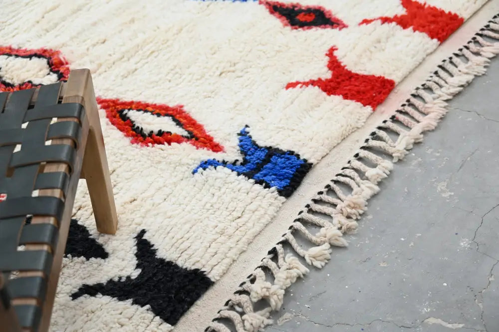 A cream-colored handmade moroccan azilal rug with bold red blue and black geometric patterns and thick fringed edges