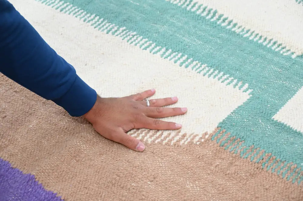 A hand with a silver ring rests on a handmade moroccan azilal rug with wide teal cream and beige stripes