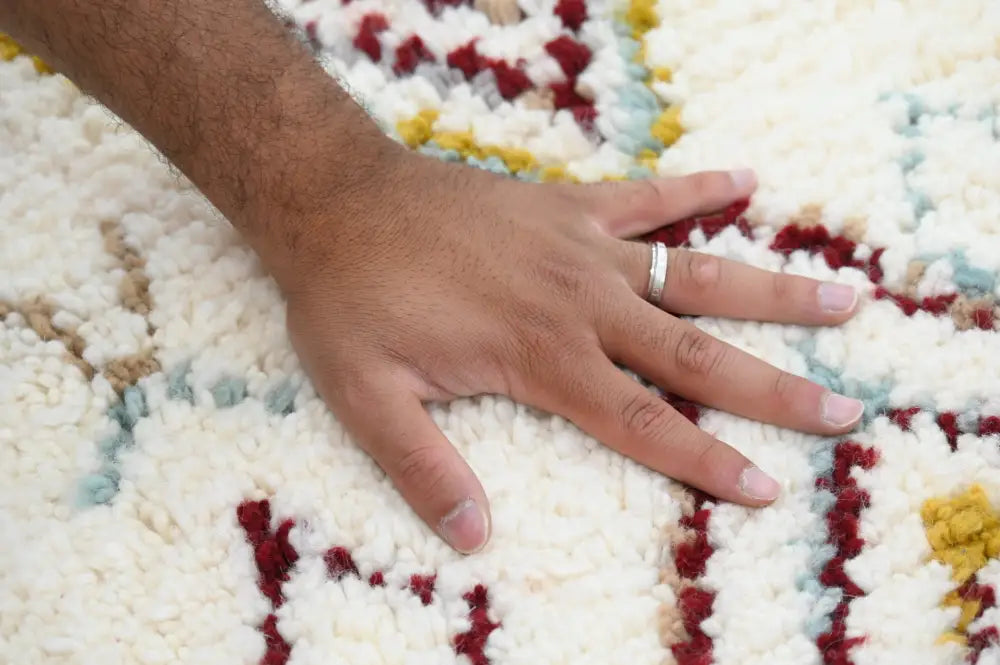 A hand with a silver ring rests on a plush cream handmade moroccan azilal rug with red and yellow geometric patterns