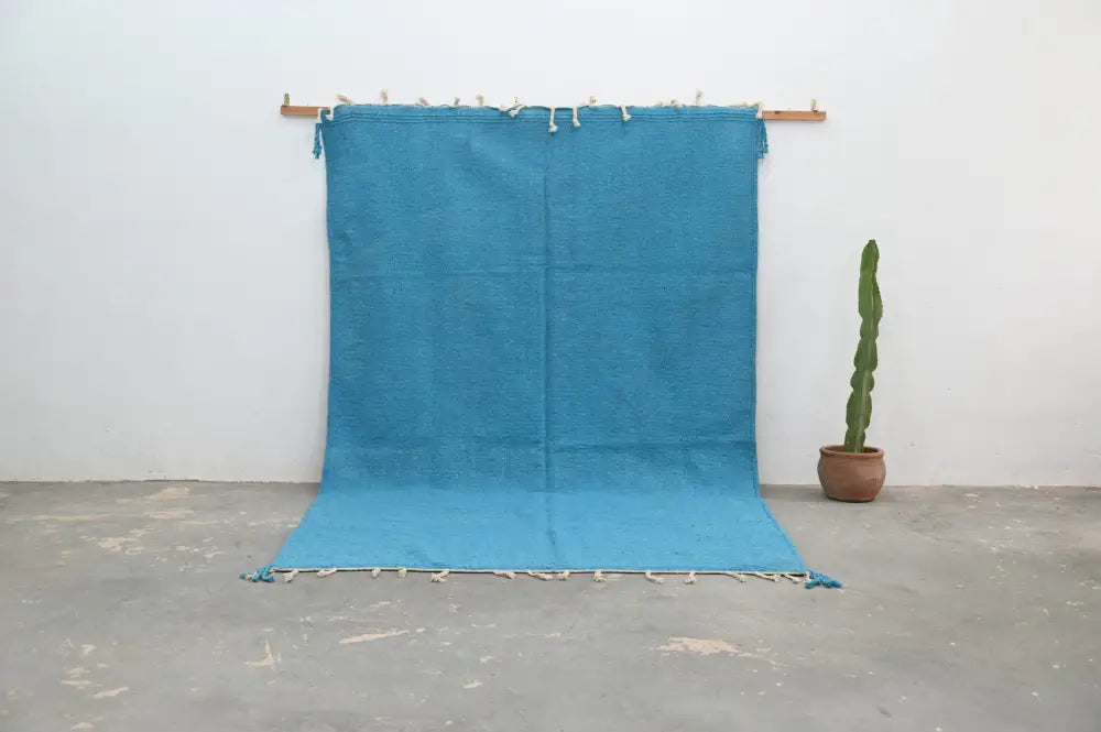 A vibrant turquoise handmade azilal rug 306x200cm with fringed edges and tassels displayed on a wooden rod